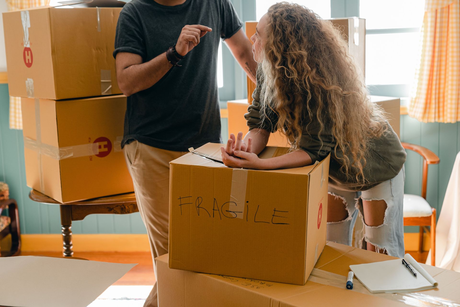 Crop faceless man in wristwatch talking to girlfriend in fashion jeans with wavy hair while standing near box with FRAGILE note after moving into new house